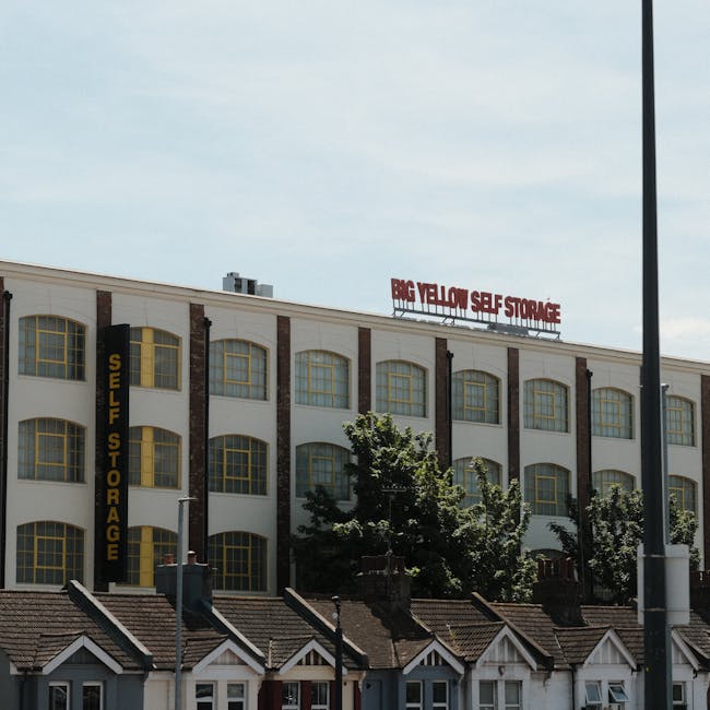 The exterior of a multi-storey building with a sign reading 'BIG YELLOW SELF STORAGE' in red letters on the roof, and additional yellow and black signage indicating 'SELF STORAGE' on the side. The building features large yellow-framed windows against a white facade with brick accents. In front of the building, a row of small, pitched-roof houses with white frames and dark roofing tiles is visible, along with some trees and shrubs partially obscuring the lower part of the structure. The scene is captured during daytime with clear skies, and a tall lamppost stands on the right side of the image. The image depicts the environment where home relocation and furniture transport services by Man With a Van Tottenham Hale might operate, emphasizing storage options and the logistics involved in apartment moves near Tottenham Hale Station.