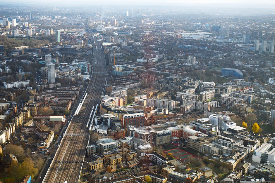 An aerial view of Tottenham Hale, London, showing a densely built urban area with numerous mid-rise residential and commercial buildings, some with flat roofs and various architectural styles. A major railway line runs through the centre of the image, with tracks visible on the left side, indicating active train movement. The surrounding streets are lined with trees, and there are several open spaces, including a sports court and park areas. The skyline extends into the distance with a mix of high-rise and low-rise structures, under a partly cloudy sky. This scene depicts typical foreground and background elements associated with city centre home relocations and furniture transport logistics, relevant to house removal services operated by companies like Man With a Van Tottenham Hale.