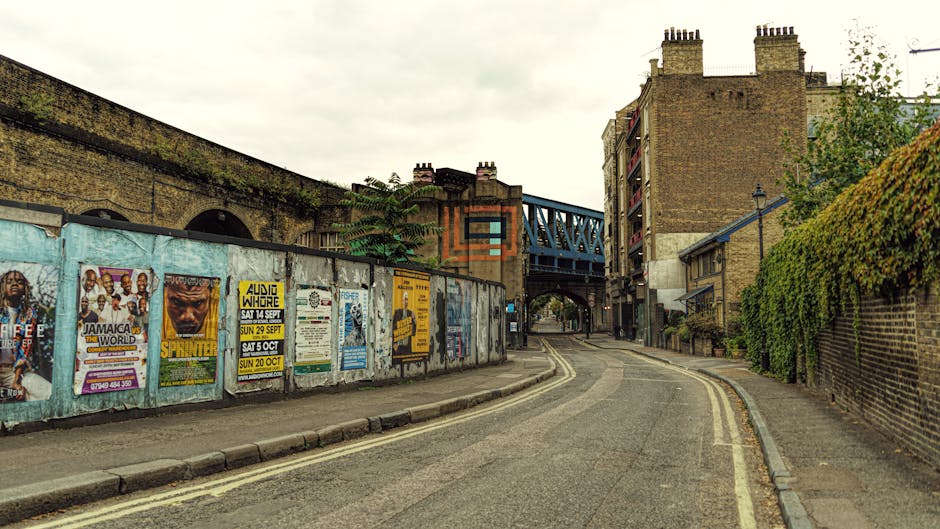 A narrow urban street scene in Tottenham Hale with a slight curve leading to an underpass and a blue railway bridge above. On the left side, a tall brick wall is decorated with colorful posters advert