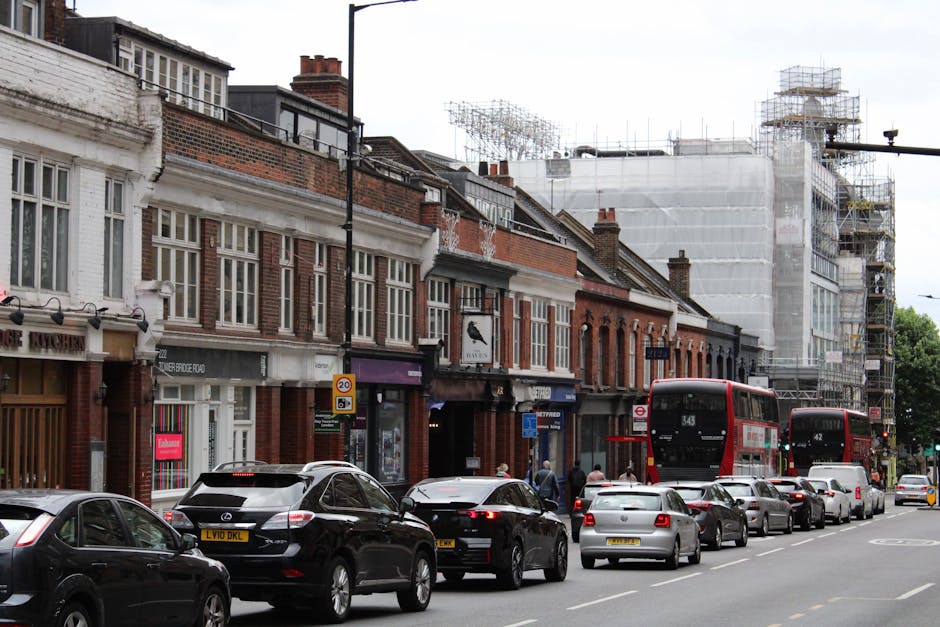 A narrow urban street scene in Tottenham Hale with a slight curve leading to an underpass and a blue railway bridge above. On the left side, a tall brick wall is decorated with colorful posters advert