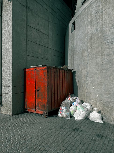 A red, weathered metal waste container situated on a dark grey tiled pavement next to a light grey concrete wall and a darker grey, smooth stone building exterior in Tottenham Hale. Several white and transparent plastic garbage bags filled with mixed household waste are piled next to the container, some leaning against it. The setting appears to be outside a property, with no visible signage. The image includes partial views of a doorway and a narrow alleyway, suggesting this is an area designated for waste disposal or clearance during home removal or moving services. Man With a Van Tottenham Hale specialises in removals and assisting with the clearance of bulky waste, as reflected in the scene showing a typical waste disposal setup during packing and furniture transport processes.