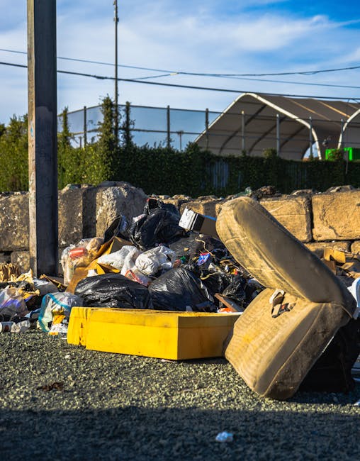 A pile of bulky waste, including a discarded sofa cushion with fabric cover and visible foam padding, several black plastic garbage bags filled with household waste, and various cardboard boxes and packaging materials, all situated on a gravel surface next to a stone wall. The waste is outside a property, with a metal pole partially visible on the left side of the image and overhead power lines running across the sky. In the background, there is a green hedge, a structure covered with a white canopy supported by metal poles, and a partly cloudy sky. This scene illustrates the process of clearing unwanted items, relevant to house removals and furniture transport services, and aligns with quick disposal options in Tottenham Hale as provided by Man With a Van Tottenham Hale.