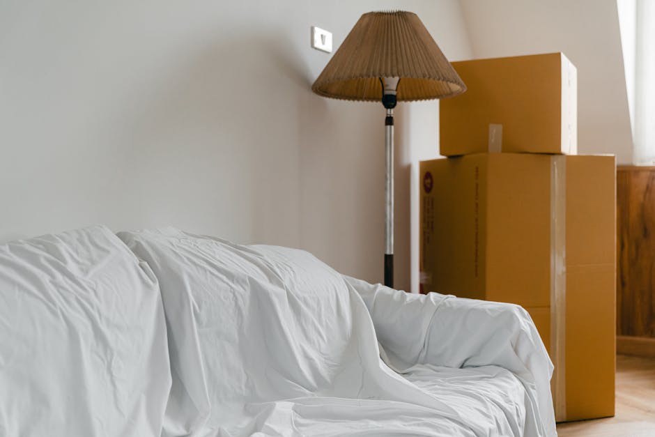 Inside a home's living room, with a white upholstered sofa covered in a white sheet or protective cover, positioned against a plain light-colored wall. To the right, there are three large cardboard boxes stacked on top of each other, secured with clear packing tape, indicative of recent packing or moving activities. A tall, beige fabric lampshade on a black metal stand is situated between the sofa and boxes, providing ambient lighting. The floor appears to be wooden or laminate, and the environment suggests an ongoing house relocation or furniture transport process facilitated by Man With a Van Tottenham Hale. The scene captures a moment during packing or unpacking, with items either prepared for removal or awaiting placement in a new property, consistent with services in removals and home moving.