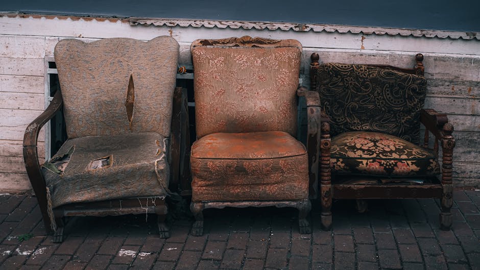 Three old, worn-out antique chairs placed outdoors against a weathered white brick wall with peeling paint. The left chair has a damaged, torn fabric upholstery with stuffing exposed and one armrest missing, revealing wooden framework. The middle chair features a faded, stained fabric with visible wear and tear on the seat and backrest, showcasing a classic design with short, curved wooden legs. The right chair is a traditional wooden armchair with patterned fabric cushions in black, gold, and red, and has turned wooden spindles on the armrests and back. These chairs are positioned on a paved surface, likely outside a property undergoing a house removal or relocation process, which is associated with services provided by Man With a Van Tottenham Hale. The scene highlights furniture transport and packing considerations during house moves.