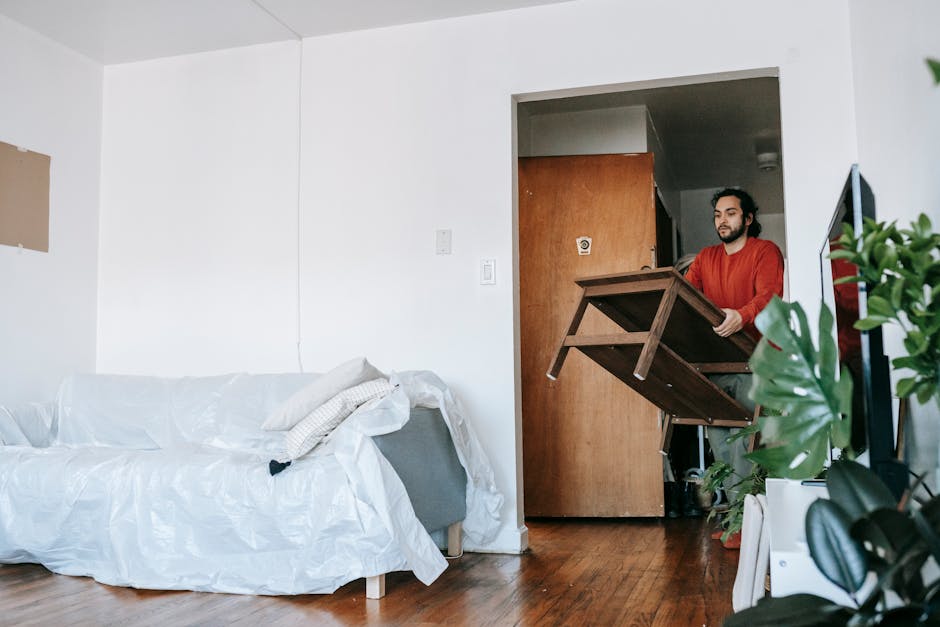 Inside a home's living room, with a white upholstered sofa covered in a white sheet or protective cover, positioned against a plain light-colored wall. To the right, there are three large cardboard boxes stacked on top of each other, secured with clear packing tape, indicative of recent packing or moving activities. A tall, beige fabric lampshade on a black metal stand is situated between the sofa and boxes, providing ambient lighting. The floor appears to be wooden or laminate, and the environment suggests an ongoing house relocation or furniture transport process facilitated by Man With a Van Tottenham Hale. The scene captures a moment during packing or unpacking, with items either prepared for removal or awaiting placement in a new property, consistent with services in removals and home moving.