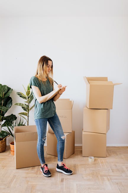 A young woman with shoulder-length blonde hair, wearing a casual green t-shirt, ripped blue jeans, and black and pink sneakers, standing inside a home surrounded by cardboard moving boxes in various sizes. She is holding a pen and a notepad, appearing to be organizing or taking inventory during a home relocation process. To her left, there is a green potted plant with large leaves placed on the floor, and the background features a plain white wall and light wooden flooring. The boxes are stacked and placed on the floor, some open with flaps folded back, indicative of packing or unpacking activities as part of furniture transport and packing and moving preparations. The scene reflects typical house removal activities as part of a professional move, with natural lighting highlighting the interior space, aligning with high-quality, factual visuals related to house removals and relocation services offered by Man With a Van Tottenham Hale.