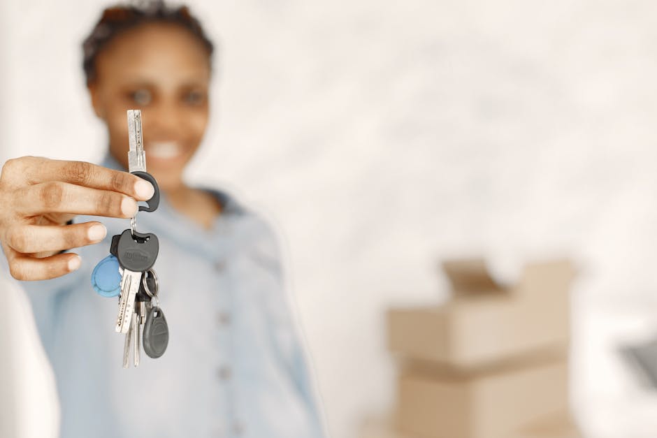 A woman with dark hair tied back is extending her hand towards the camera, holding a set of keys with multiple keychains attached. She is smiling and dressed in a light blue shirt, standing indoors with a blurred background. Behind her, there are several cardboard moving boxes stacked on top of each other, some open with packing materials visible. The scene appears to be inside a property ready for a home relocation or furniture transport, with natural lighting illuminating the area. The focus is on the keys, signifying the completion of a move or the handing over of property. This image aligns with the context of house removals and packing and moving services provided by Man With a Van Tottenham Hale, supporting the process of interior packing, loading, and transport within the relocation logistics.