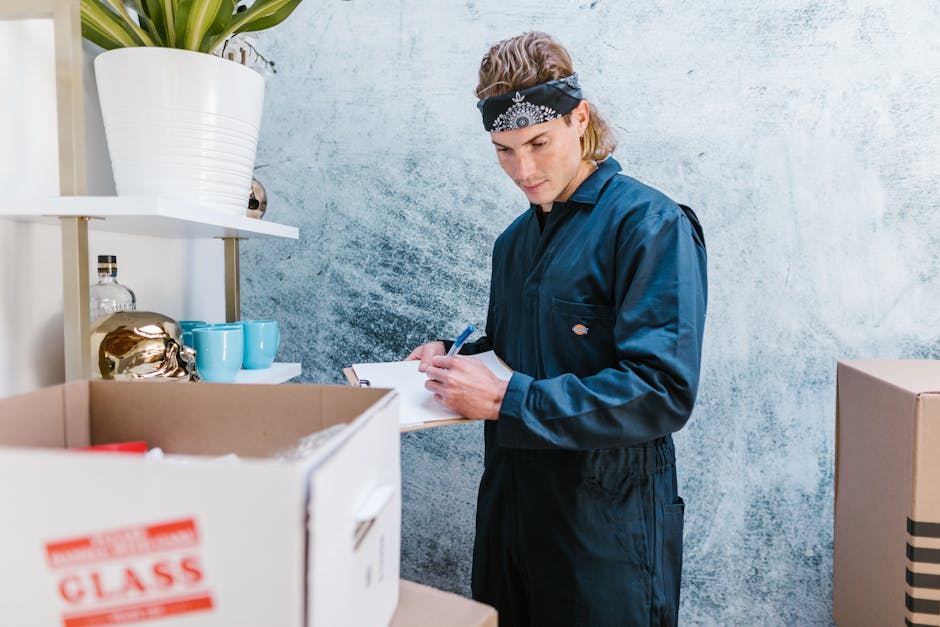 A young woman with shoulder-length blonde hair, wearing a casual green t-shirt, ripped blue jeans, and black and pink sneakers, standing inside a home surrounded by cardboard moving boxes in various sizes. She is holding a pen and a notepad, appearing to be organizing or taking inventory during a home relocation process. To her left, there is a green potted plant with large leaves placed on the floor, and the background features a plain white wall and light wooden flooring. The boxes are stacked and placed on the floor, some open with flaps folded back, indicative of packing or unpacking activities as part of furniture transport and packing and moving preparations. The scene reflects typical house removal activities as part of a professional move, with natural lighting highlighting the interior space, aligning with high-quality, factual visuals related to house removals and relocation services offered by Man With a Van Tottenham Hale.