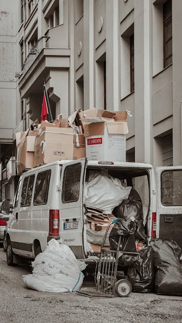 A white commercial van parked on a street in an urban area with multi-storey residential buildings. The van's rear doors are open, revealing an interior filled with large black plastic garbage bags, rolls of packing materials, and cardboard boxes. Several packed cardboard boxes, some open and others sealed, are stacked on top of the van, secured with plastic wrap and packing tape. A small trolley with a few cardboard pieces sits on the pavement next to the van, indicating ongoing loading or unloading activity. The scene reflects the home relocation process, with furniture and belongings prepared for transport by a professional removals service such as Man With a Van Tottenham Hale, supporting efficient furniture transport and packing and moving operations. The environment is well-lit with natural daylight, and the setting suggests an urban street ready for a house move compliant with local council permit rules for moving vans in N17, Tottenham Hale.