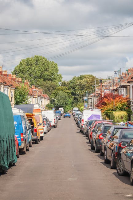 A residential street scene showing tightly parked cars lining both sides of the asphalt road, with some vehicles covered by protective tarps or fitted with roof racks. The street is bordered by terraced houses with brick facades, small front gardens, and some with colorful foliage trees, indicating a suburban neighbourhood. Overhead, power lines run across the sky, which is partly cloudy with patches of blue. In the distance, a narrow lane extends beyond the parked vehicles, with more greenery and larger trees visible further back. The setting appears suitable for a home relocation or furniture transport operation, typical of the planning involved in house removals, as demonstrated by local parking regulations often requiring permits in areas like Tottenham Hale. This image, associated with Man With a Van Tottenham Hale, illustrates an urban environment where careful loading and unloading of household items, including cardboard boxes, wooden furniture, packaging materials, and appliances, typically occurs during the process of packing and moving services.