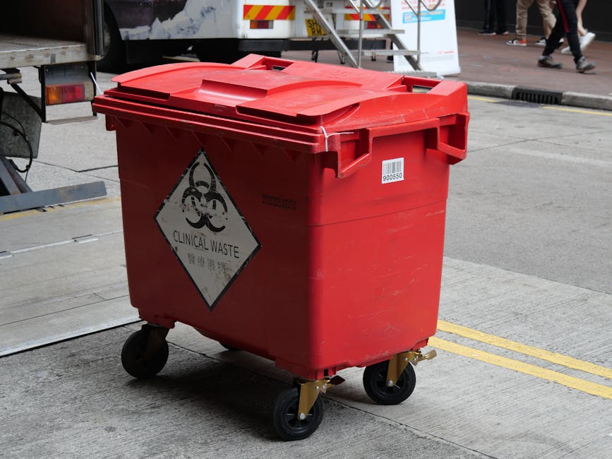 A red wheeled container with a hinged lid, marked with a hazardous waste symbol and labeled 'Clinical Waste,' positioned on a paved area next to a delivery vehicle. The container is situated outside a building door, with a metal ramp and loading equipment visible nearby. In the background, pedestrians are walking on the pavement, and the area appears to be part of an urban sidewalk or loading bay. The container is used for collecting and transporting medical or hazardous waste, which is relevant to house removals or relocation services when handling sensitive or hazardous materials. This image highlights the importance of safety and proper disposal procedures during a home relocation, exemplifying how waste items are managed in the moving process and the role of specialist removal companies like Man With a Van Tottenham Hale.