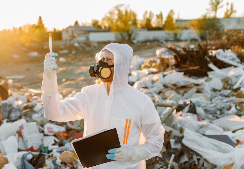 A person wearing protective gear, including a white disposable coverall, a black gas mask with yellow filters, safety goggles, and white gloves, holding a small orange and white stick in one hand and a clipboard in the other, standing amidst a large, open outdoor area filled with scattered household waste and debris. The background features piles of discarded items such as cardboard boxes, plastic containers, and other rubbish, with some structures and trees visible in the distance under a clear sky. The scene appears to be during late afternoon or early evening, with warm sunlight illuminating the surroundings. This image illustrates the hazardous waste management process involved in house relocation or furniture transport, highlighting safety precautions taken by Man With a Van Tottenham Hale during waste removal and packing of items for a home move, ensuring compliance with hazardous waste rules for movers in Tottenham Hale.