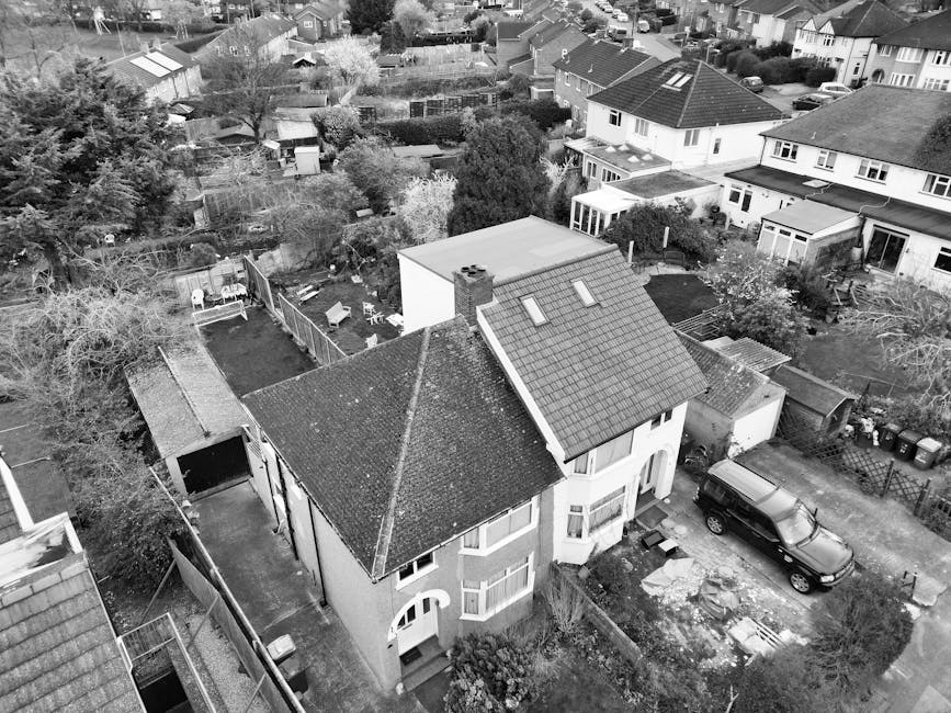 An aerial black-and-white photograph of a residential property in Tottenham Hale, showing a two-storey house with a pitched roof, dhidden skylights, and a small front garden. The house is situated on a paved driveway with a dark SUV parked nearby. The property is surrounded by a mix of other houses, some with gardens, fences, and outbuildings. Visible elements include cardboard boxes and packing materials on the driveway, indicating ongoing packing or disassembly as part of a home relocation process. The backyard is enclosed by a wooden fence and contains garden furniture, trees, and a small shed. The scene captures the outdoor environment during daylight, with surrounding trees and neighboring homes in the background, providing a contextual view of a typical house move setup in Tottenham Hale, supported by the services of Man With a Van Tottenham Hale for furniture transport and house removals.