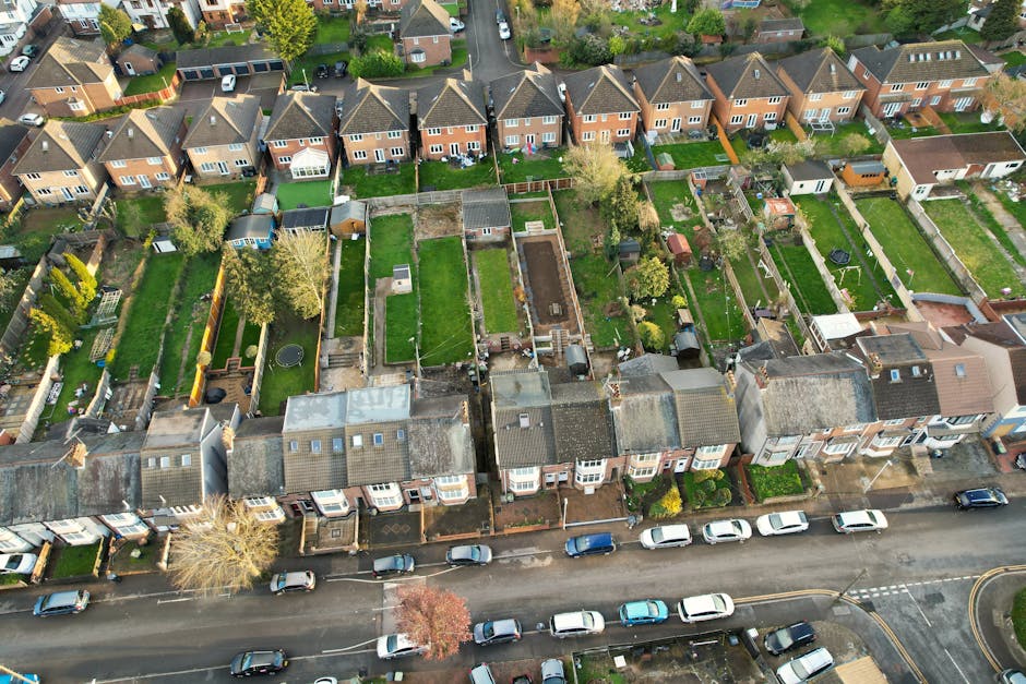 An aerial view of a residential street showing a row of terraced houses at the front with pitched roofs, and numerous back gardens with fences, lawn areas, sheds, and outdoor furniture. Visible in the foreground are parked cars along the street and a sidewalk. In some gardens, there are items such as garden furniture, toys, and equipment, with one garden featuring a trampoline. The image captures the outdoor environment in daylight, with clear weather, highlighting the typical layout of houses and their private yard spaces, suitable for a home relocation or furniture transport process involving a removal service such as Man With a Van Tottenham Hale.