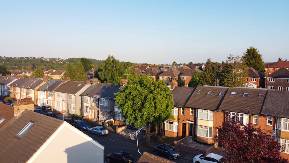 An aerial black-and-white photograph of a residential property in Tottenham Hale, showing a two-storey house with a pitched roof, dhidden skylights, and a small front garden. The house is situated on a paved driveway with a dark SUV parked nearby. The property is surrounded by a mix of other houses, some with gardens, fences, and outbuildings. Visible elements include cardboard boxes and packing materials on the driveway, indicating ongoing packing or disassembly as part of a home relocation process. The backyard is enclosed by a wooden fence and contains garden furniture, trees, and a small shed. The scene captures the outdoor environment during daylight, with surrounding trees and neighboring homes in the background, providing a contextual view of a typical house move setup in Tottenham Hale, supported by the services of Man With a Van Tottenham Hale for furniture transport and house removals.
