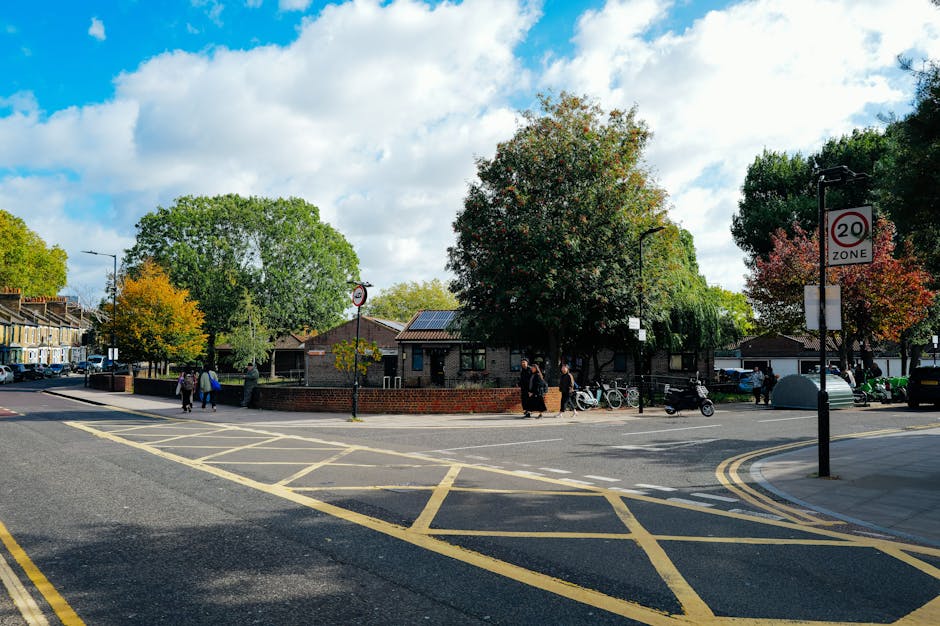 A wide street scene in Broadwater Farm, Tottenham Hale, showing a road with yellow box junction markings and a 20 mph speed zone sign. Several pedestrians are walking along the sidewalk, some near bicycles and a motorcycle parked by a low brick wall. There are trees with green and autumn-colored leaves lining the street. In the background, a single-story building with a dark roof is partially visible behind the foliage. The sky is partly cloudy with patches of blue, and the scene appears to be during daylight. This setting illustrates a typical residential area where home relocation and furniture transport might take place, supported by nearby parking spaces and open space suitable for loading and unloading during house removals, which Man With a Van Tottenham Hale could assist with.
