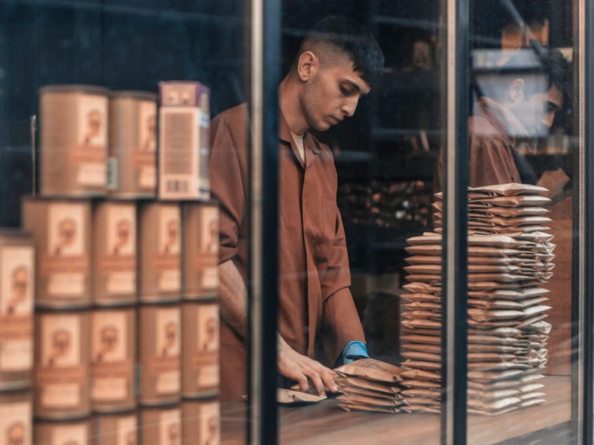 A man wearing a brown shirt and blue gloves is inside a storage room, visible through a glass window or door, carefully folding or sealing a cardboard package. To his left, there is a stack of similarly packed cardboard boxes with printed labels, and to his right, a tall organized stack of padded envelopes or parcels, likely prepared for a furniture transport or packing process. The environment appears to be well-lit, with a focus on meticulous packing, reflecting the careful handling associated with home relocation or business removals at Riverside Retail Park. The scene captures a moment of packing and moving logistics, as part of the services provided by Man With a Van Tottenham Hale, specializing in removals and relocation services. The overall setting suggests a systematic and professional approach to packing and moving tasks, supporting an efficient loading process into a vehicle or storage area.
