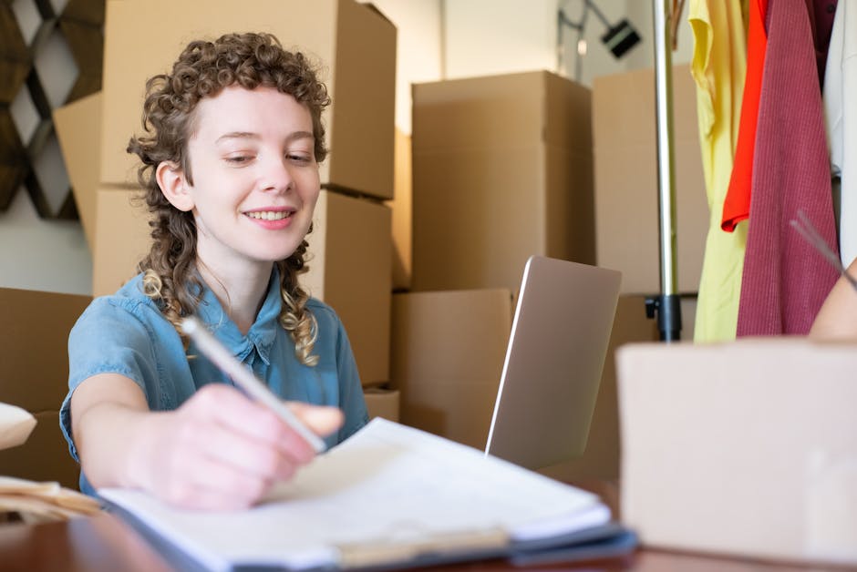 A woman with curly hair, dressed in a light blue shirt, is seated at a desk surrounded by moving and packing materials, including cardboard boxes and a laptop. She is smiling slightly and appears to be writing or taking notes with a pen. Behind her, stacked cardboard boxes of various sizes are placed in a room used for packing items before a house or office move. To her right, a clothing rack with hanging garments and a tripod with a light are visible, indicating an environment prepared for packing and relocating belongings. The setting has warm lighting and a tidy, organized appearance, reflecting a professional packing and moving process undertaken by Man With a Van Tottenham Hale as part of a furniture transport and home relocation service, supporting the planning and logistics of house removals and packing strategies for businesses at Riverside Retail Park in Tottenham Hale.
