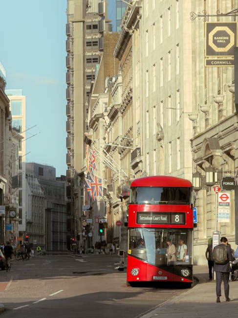 A red double-decker bus on a busy city street surrounded by tall, historic buildings with classic architecture, including decorative cornices and ornate window frames. The bus displays route number 8 and is heading towards Tottenham Court Road. Pedestrians are walking along the sidewalk, some carrying backpacks or shopping bags. Shop signs and streetlights are visible, along with a British flag hanging between buildings. The scene is well-lit with daylight, capturing the urban environment typical of central London, near areas like Tottenham Hale. This setting is relevant for house removals and furniture transport as it depicts the streets where relocation vehicles, such as vans or trucks from Man With a Van Tottenham Hale, might operate during home relocation or packing and moving processes.