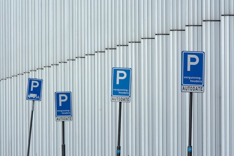 Four blue parking restriction signs with white 'P' symbols and text indicating parking permissions and permit validation are mounted on black metal posts against a vertically ribbed metal wall with a light gray finish. The signs display information about parking permits and the need for automatic validation, with two of them showing small illustrations of trucks. The signs are arranged in a line, with varying heights, on a paved area or sidewalk near a building entrance, suggesting an urban setting related to parking management. The scene is well-lit with natural light, and the positioning of the signs supports understanding of parking regulations near High Rd Tottenham, relevant to local house removals and transportation services managed by Man With a Van Tottenham Hale.