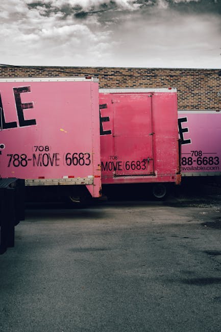 A row of three large pink moving containers with black and purple text displaying contact information, positioned on the side of a paved street next to a brick wall. The containers are made of metal a