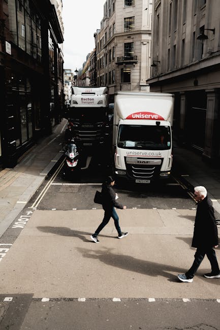 A view of a narrow urban street showing a white moving truck with the logo 'uniserve' parked partially on the sidewalk, along with a smaller black vehicle parked beside it. In the foreground, two pedestrians—a woman carrying a black bag and a man with white hair—are walking across a pedestrian crossing on the street. On the left side, there are black metal balconies attached to commercial buildings with large windows, while on the right, multi-storey buildings with traditional architecture are visible. The scene depicts typical elements of home relocation logistics, with vehicles positioned for furniture transport and moving supplies, and the pedestrians involved in routine street activity during a moving operation. The overcast sky provides diffuse lighting across the scene.