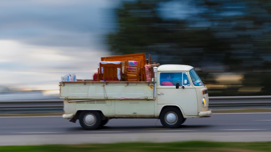 A vintage cream-colored cargo van with a compact and rounded design is driving along a motorway during daytime, with motion blur indicating high speed. The van's flatbed cargo area is loaded with various household items prepared for a home relocation, including a small wooden dining table with chairs, wrapped in protective plastic, along with cardboard boxes, plastic bags, and packing materials. The interior visible through the windshield shows a pink object on the dashboard. The surrounding environment features blurred trees and a cloudy sky, emphasizing the vehicle's movement. Man With a Van Tottenham Hale, a professional removals service, is involved in this furniture transport process, supporting efficient packing and loading during house removals, as reflected in the scene's focus on packing and transport logistics.