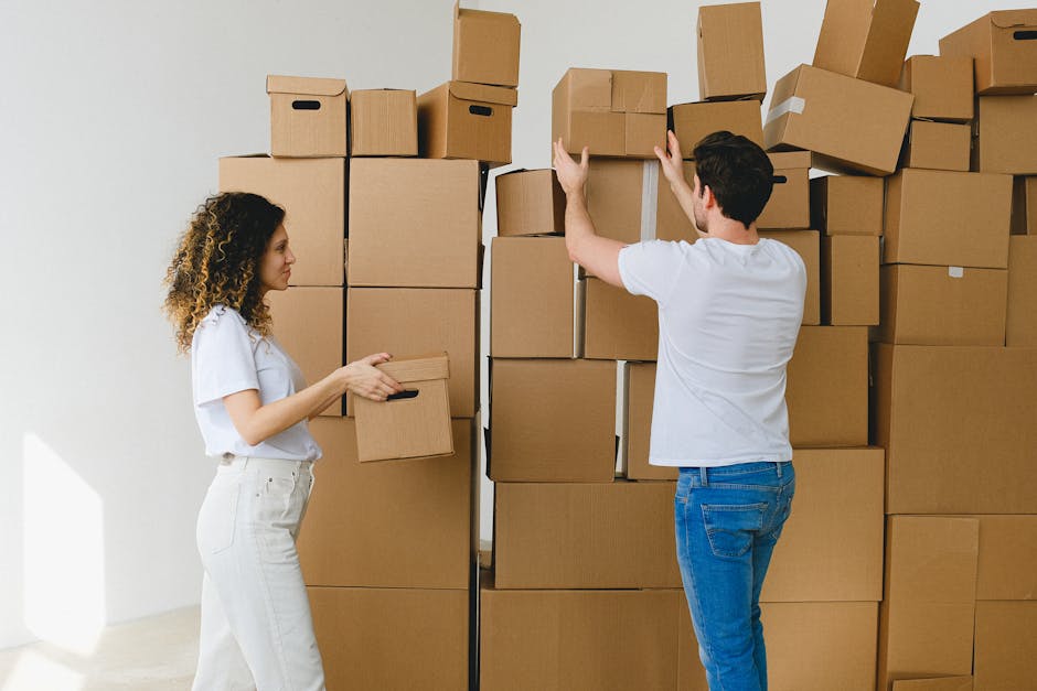 Two movers from Man With a Van Tottenham Hale are indoors, handling packing and loading materials for a home relocation. The man, wearing a white t-shirt and blue jeans, is lifting a cardboard box towards a stack of similar boxes arranged in an organized manner against a plain white wall. The woman, dressed in a white top and light-colored pants, is holding a smaller cardboard box, preparing to place it on or move it from the stack. The scene includes a variety of cardboard boxes of different sizes, some with white packing tape, positioned on the floor and stacked vertically, indicating the packing process for furniture transport or moving services. The bright lighting highlights the indoor environment, suggesting a professional moving operation involving meticulous packing and careful stacking, typical of house removals or last-minute moving help, as offered by [COMPANY_NAME].