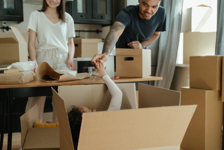 A man and a woman are inside a room surrounded by moving boxes, with the man leaning over a cardboard box, handing a glass of water to a child sitting inside the box. The woman stands nearby, watching the interaction with a slight smile. The room contains several packed cardboard boxes of various sizes, some with packing paper and bubble wrap visible. The man is wearing a dark t-shirt, and the woman is dressed in a light-colored top and trousers. Natural light enters through a window with curtains on the right side of the image. The scene depicts a home relocation process involving packing and preparation for moving, with the furniture and boxes arranged in the room as part of an ongoing moving or packing activity, which [COMPANY_NAME] can assist with during last-minute removals in N17, Tottenham Hale.