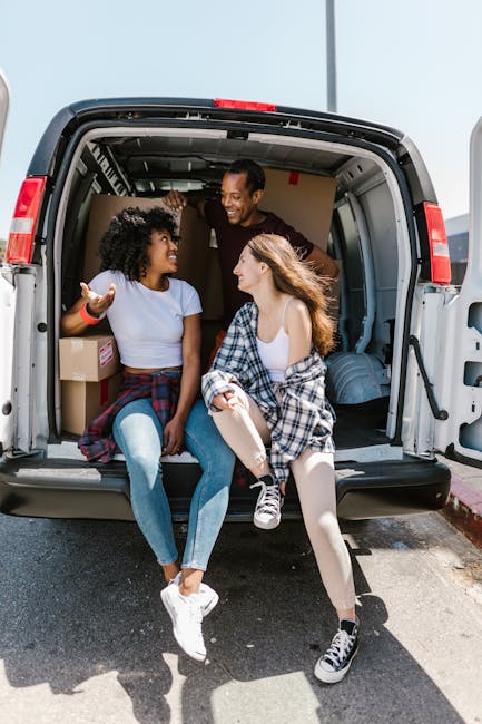 Two movers from Man With a Van Tottenham Hale are indoors, handling packing and loading materials for a home relocation. The man, wearing a white t-shirt and blue jeans, is lifting a cardboard box towards a stack of similar boxes arranged in an organized manner against a plain white wall. The woman, dressed in a white top and light-colored pants, is holding a smaller cardboard box, preparing to place it on or move it from the stack. The scene includes a variety of cardboard boxes of different sizes, some with white packing tape, positioned on the floor and stacked vertically, indicating the packing process for furniture transport or moving services. The bright lighting highlights the indoor environment, suggesting a professional moving operation involving meticulous packing and careful stacking, typical of house removals or last-minute moving help, as offered by [COMPANY_NAME].